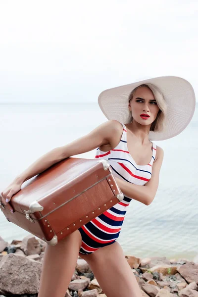 stylish tourist in swimsuit with vintage travel bag near the sea