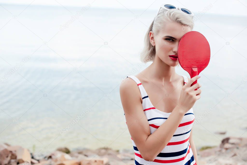 attractive sportswoman in retro striped swimsuit posing with ping pong racket on rocky beach