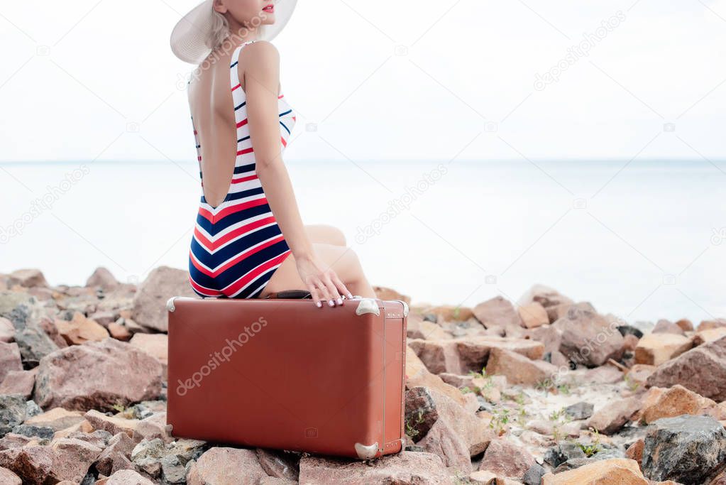 cropped view of stylish woman in striped swimsuit sitting on luggage on rocky beach