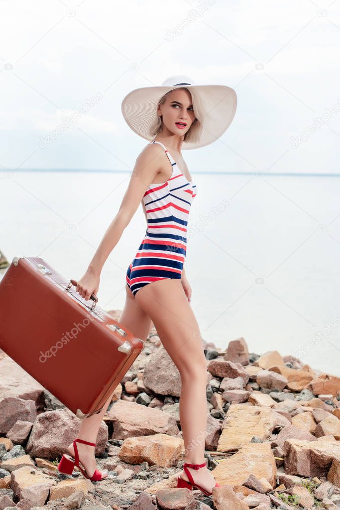 stylish womna in striped swimsuit and hat with vintage suitcase on rocky beach