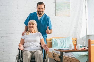 Tekerlekli sandalyeye mahkum kamera ve başparmak gösterilmesini neşeli genç doktor gülümseyen kadın kıdemli mutlu