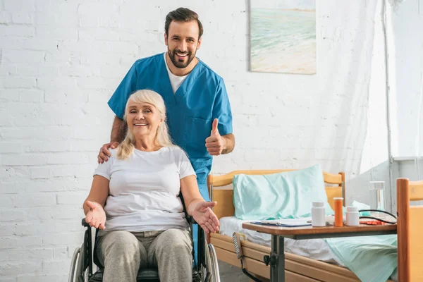 Feliz Anciana Silla Ruedas Sonriendo Cámara Alegre Joven Médico Mostrando — Foto de Stock