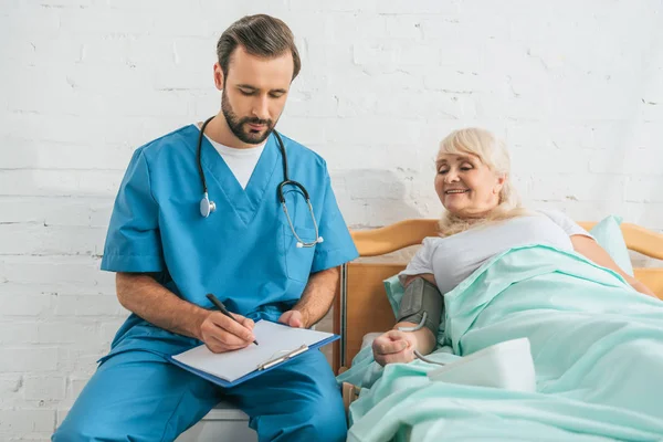 男性の看護師がベッドで横になっている笑顔の年配の女性に血圧を測定しながらクリップボードに書き込み — ストック写真