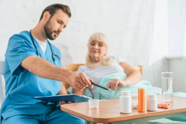 Enfermeiro Segurando Prancheta Apontando Para Pílulas Para Mulher Idosa Deitada — Fotografia de Stock