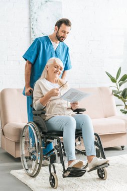 Erkek hemşire gülümseyen kadın kıdemli okuma gazete tekerlekli sandalye itmek