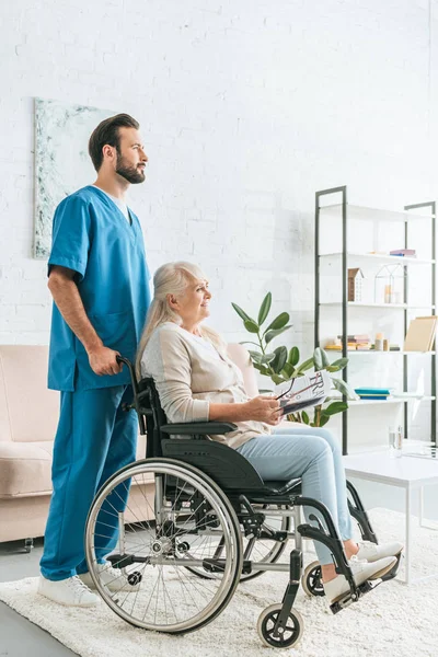 side view of social worker pushing wheelchair with senior woman holding newspaper and eyeglasses