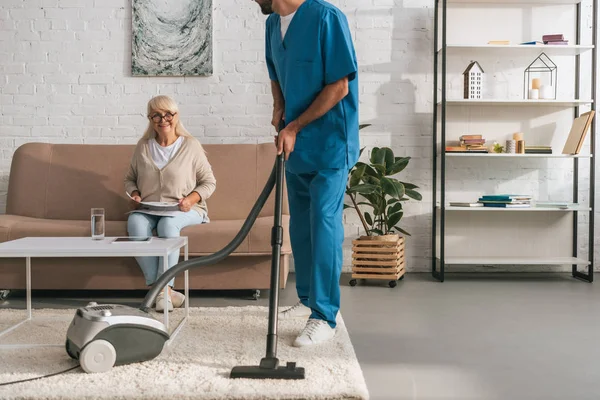 Schnappschuss Von Sozialarbeiterin Die Teppich Mit Staubsauger Reinigt Während Seniorin — Stockfoto