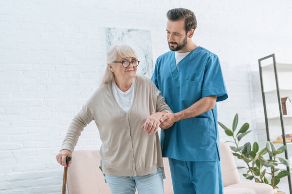social worker supporting senior woman in eyeglasses with walking stick 