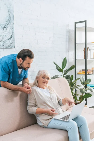 Krankenschwester Lehnt Sofa Und Schaut Seniorin Mit Laptop — kostenloses Stockfoto