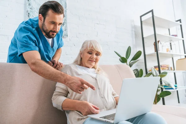 young male social worker pointing with finger at laptop white senior woman sitting on couch