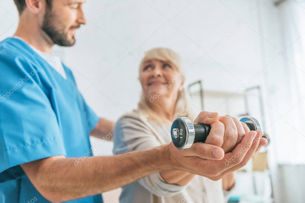 close-up view of social worker helping smiling senior woman exercising with dumbbells