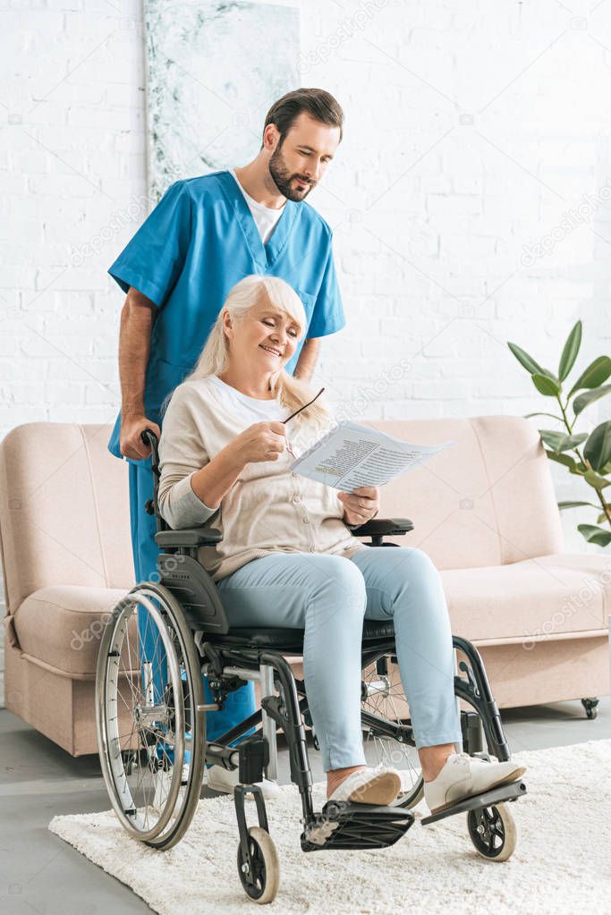 male nurse pushing wheelchair with smiling senior woman reading newspaper