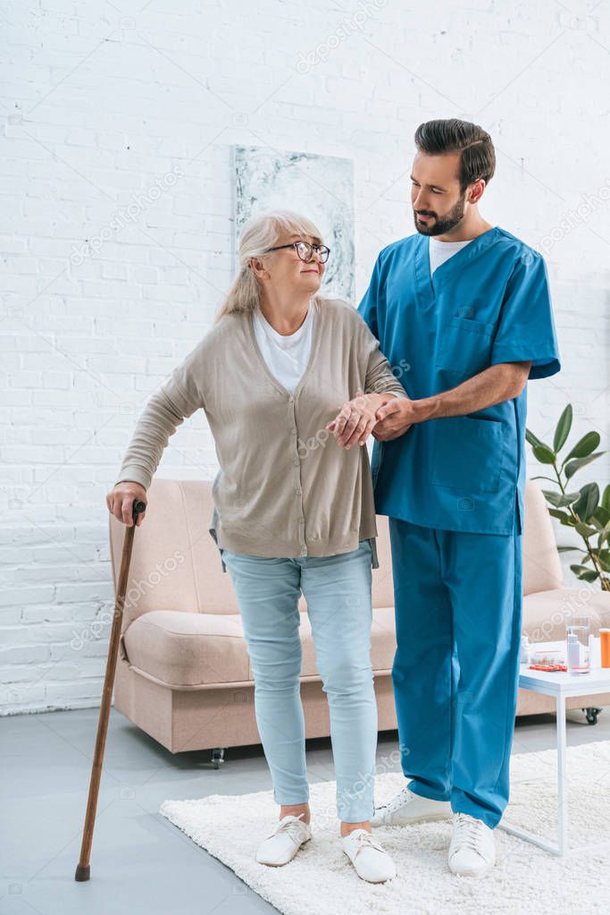 full length view of male nurse supporting senior woman in eyeglasses with walking stick 