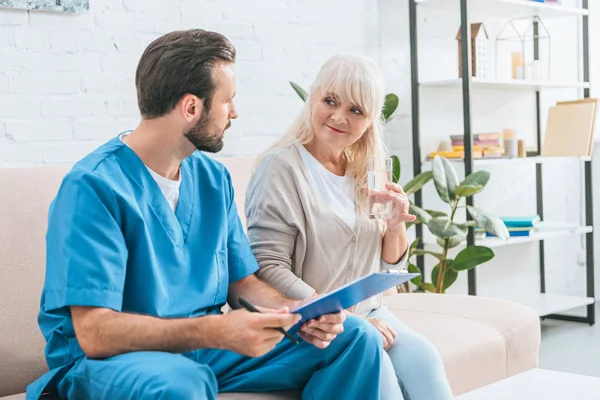 Junge Männliche Krankenschwester Hält Klemmbrett Und Blickt Lächelnde Seniorin — Stockfoto