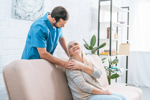 счастливая пожилая женщина сидит на диване и смотрит на молодого социального работника
 