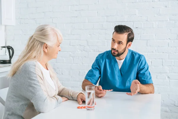 Junge Männliche Krankenschwester Hält Tabletten Und Schaut Seniorin — Stockfoto