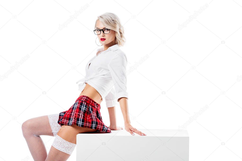 seductive woman in white shirt and plaid schoolgirl skirt sitting on white cube isolated on white