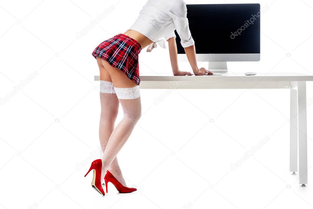 cropped shot of sexy schoolgirl in plaid skirt and stockings standing at table with blank computer screen isolated on white