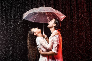 side view of romantic couple in white shirts with umbrella standing under rain on black backdrop clipart