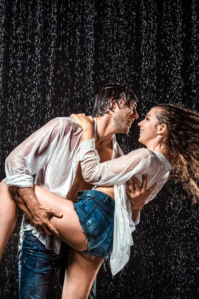 happy wet couple in white shirts standing under rain isolated on black