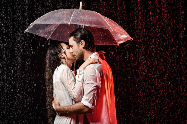 side view of romantic couple in white shirts with umbrella standing under rain on black backdrop