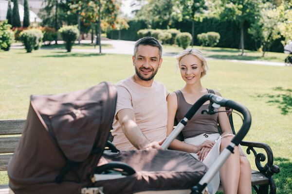 happy parents sitting on bench near baby carriage in park