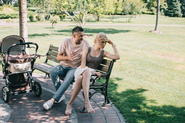 husband yelling at wife and sitting on bench near baby carriage in park 