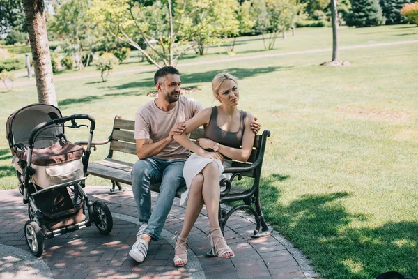 husband hugging angry wife on bench near baby carriage in park