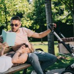 Esposa deitada nas pernas do marido perto da carruagem do bebê no parque e segurando livro, marido lendo para ela