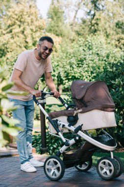 smiling father walking with baby carriage in park and looking at camera clipart