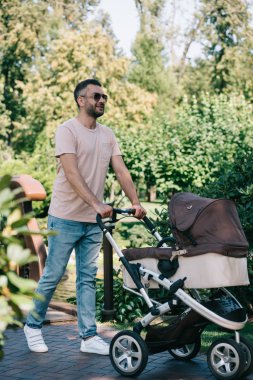 father walking with baby carriage in park clipart
