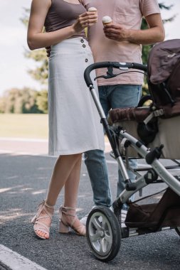 cropped image of parents holding ice cream and standing near baby carriage in park  clipart