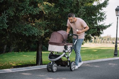 smiling father walking with baby carriage and ice cream in park and looking at baby clipart