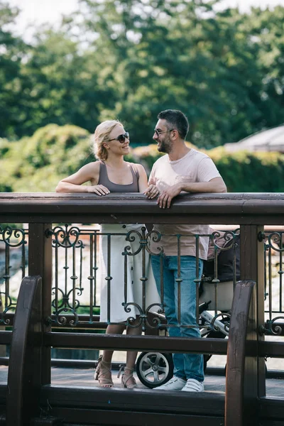 Happy Parents Standing Baby Carriage Bridge Park Looking Each Other — Free Stock Photo