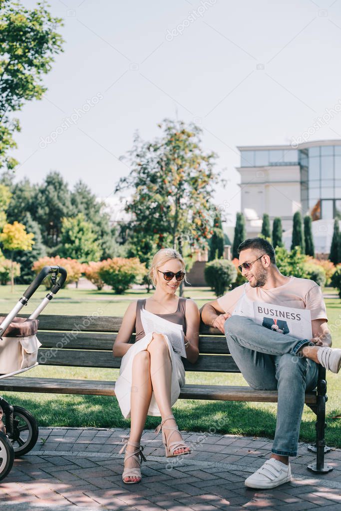 parents sitting on bench near baby carriage in park and reading magazine and business newspaper