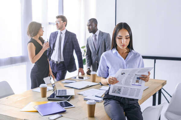 selective focus of asian businesswoman reading newspaper and colleagues having conversation behind in office