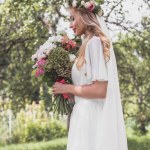 Vue latérale de jeune mariée souriante tenant bouquet de mariage dans le parc