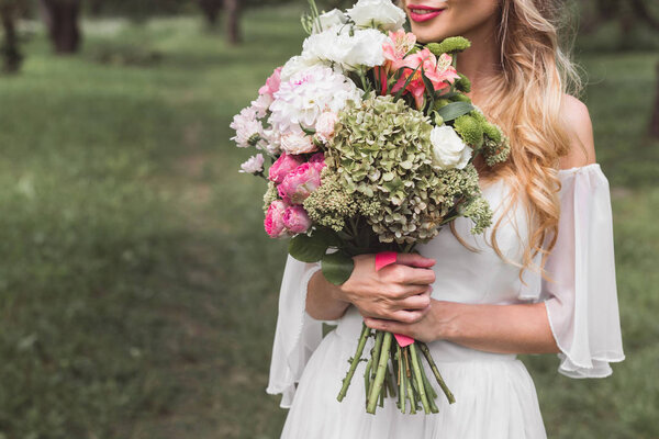 cropped shot of tender smiling blonde bride holding wedding bouquet outdoors