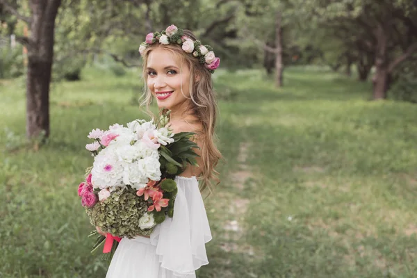 フローラル リースと花の花束を持って 屋外カメラで笑顔のウェディング ドレスの柔らかい若い花嫁 — ストック写真