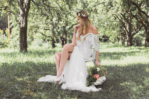 Pensive young blonde bride holding wedding bouquet and sitting on chair outdoors
