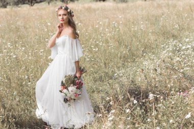 beautiful pensive young bride in wedding dress and floral wreath holding bouquet of flowers while standing outdoors clipart