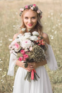 buket çiçek tutan ve kamera açık havada gülümseyen güzel mutlu genç gelin 