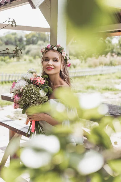 美丽的年轻新娘在花花圈的选择焦点举行婚礼花束和微笑在照相机 — 免费的图库照片