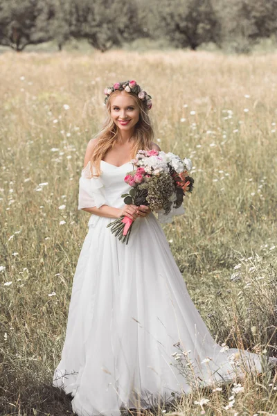 美しい若い花嫁の花束を押しながら咲くフィールドに立っている間カメラに笑顔のハイアングル  — 無料ストックフォト