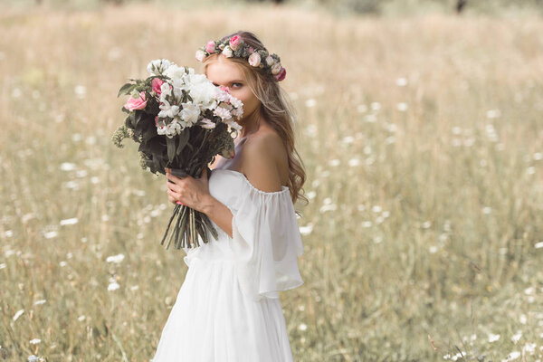 Beautiful young bride in floral wreath holding wedding bouquet and looking at camera outdoors