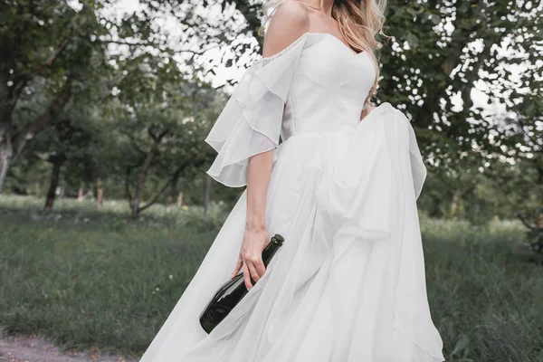 ワインのボトルを押しながら公園を歩いてのウェディング ドレスの若い花嫁のクロップ撮影  — 無料ストックフォト