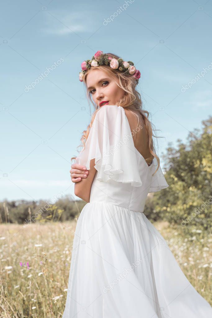tender young bride in wedding dress and floral wreath standing on field and looking at camera