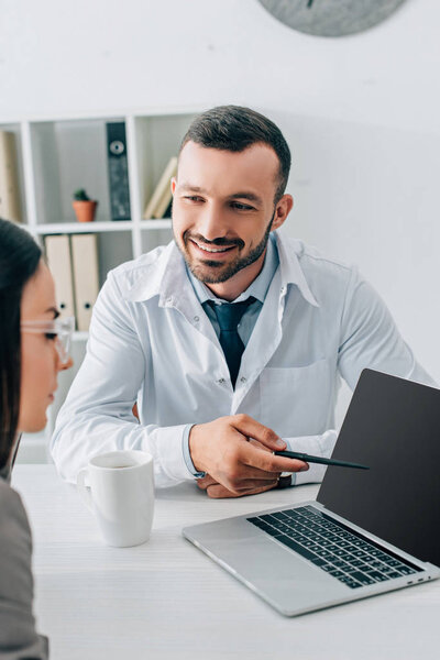smiling doctor pointing on laptop with blank screen to patient in clinic