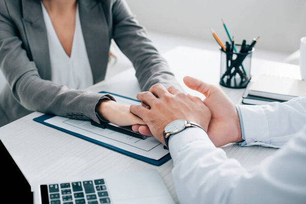 cropped image of patient and general practitioner holding hands above insurance claim form in clinic