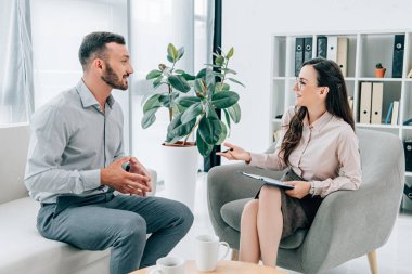 smiling psychiatrist with clipboard talking with happy patient in office clipart